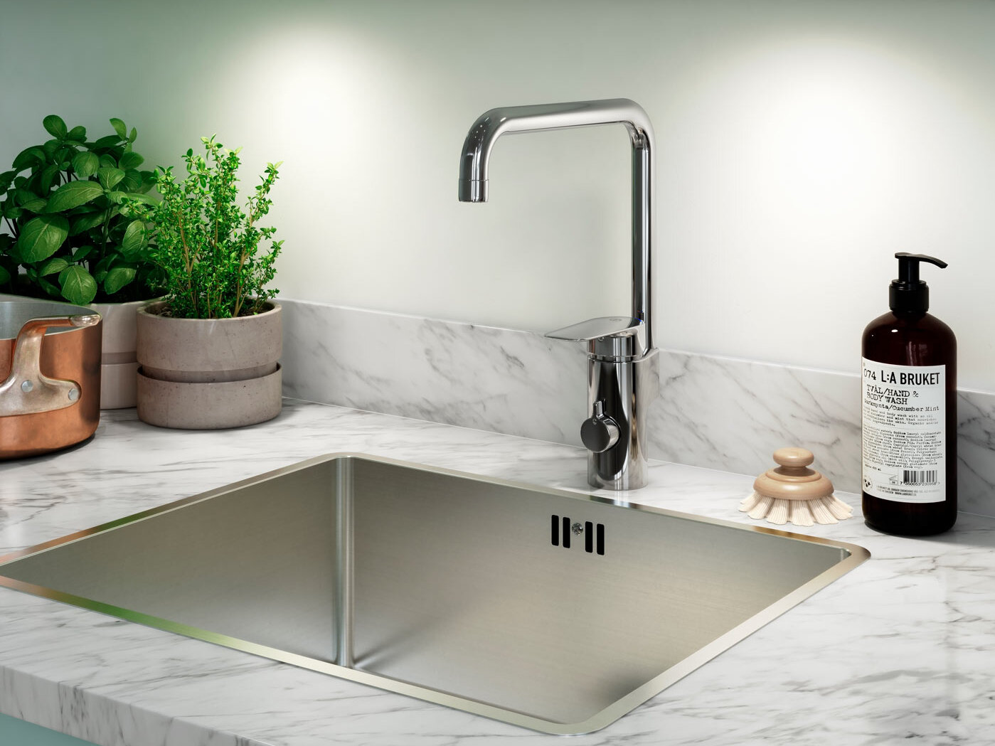Köksblandare i krom ovanför en disko i rostfrittstål, krukväxter, tvålpump och diskborste på en marmorbänk.