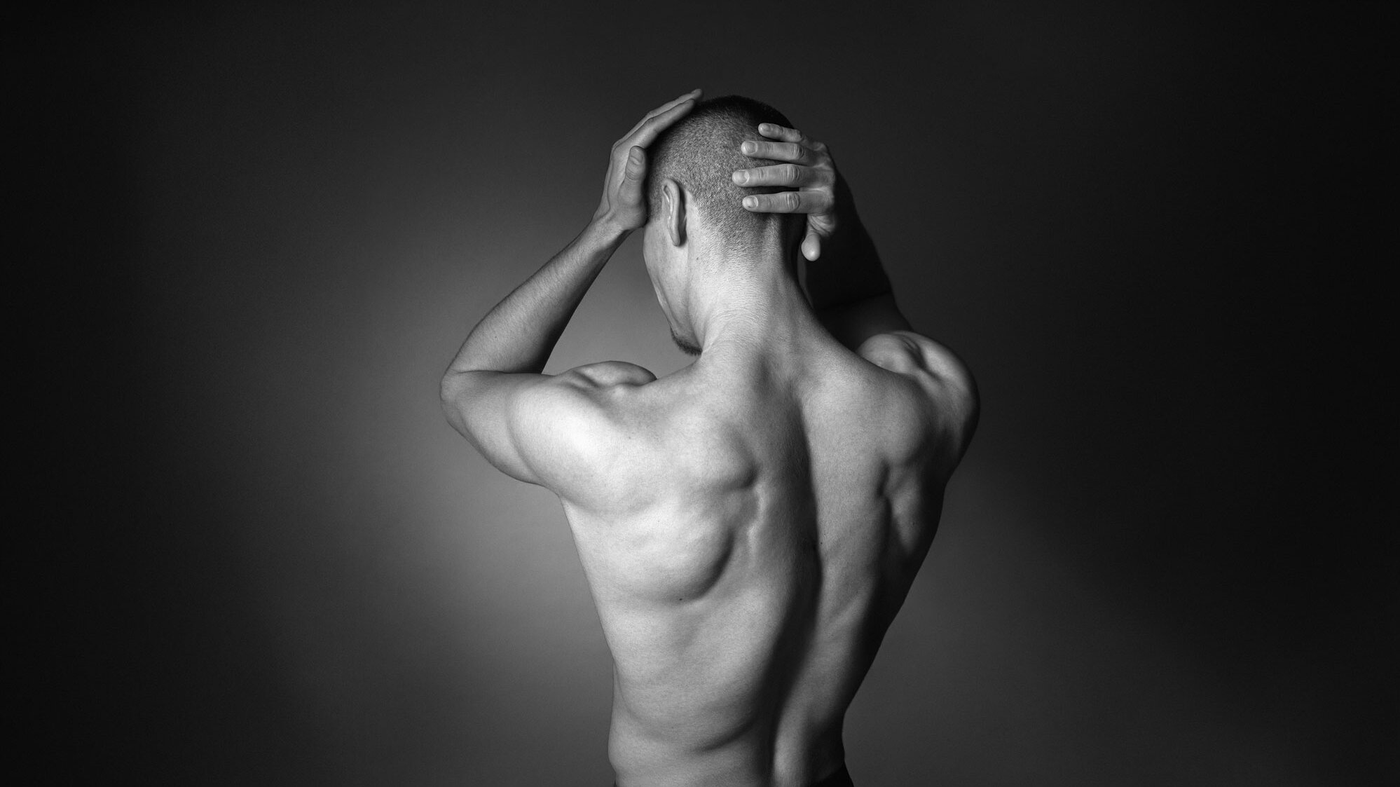 Svartvitt foto av en person sedd bakifrån, som håller huvudet med båda händerna mot en mörk bakgrund. Ser ut som att personen duschar.