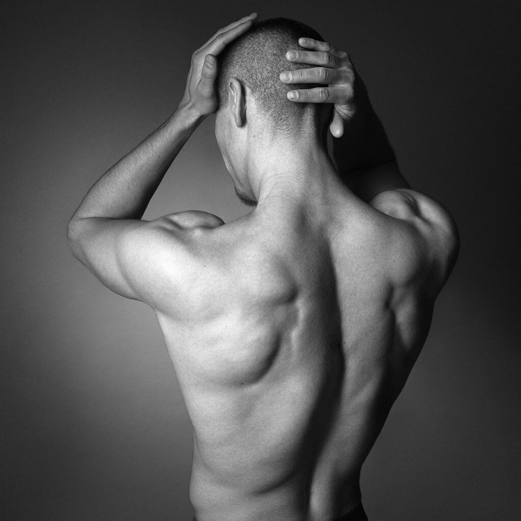 Black and white photo of a person seen from behind, holding their head with both hands against a dark background. Should be seen as taking a shower.