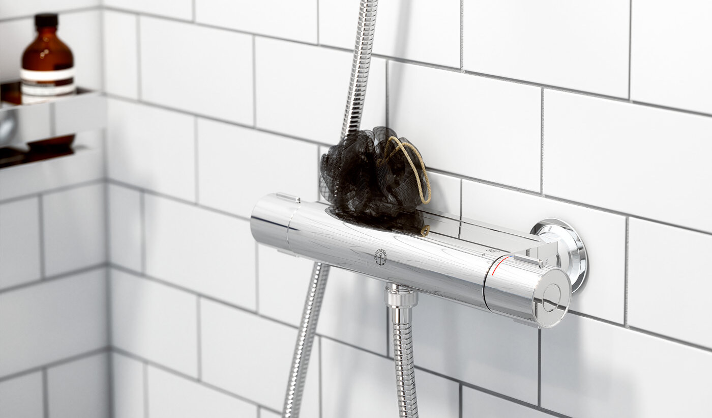Chrome Nautic shower mixer, black bath pouf on a white tiled wall; toiletries on a shelf in the background.