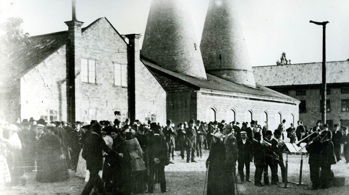 Juodai baltas vaizdas, kuriame pavaizduota grupė žmonių, švenčiančių Jonines prie Gustavsbergo mūrinių pastatų su dviem butelio formos porceliano gamybos kaminais.