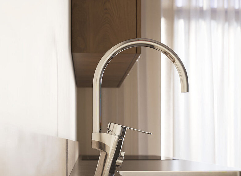 Close-up of a modern kitchen sink with a tall chrome kitchen mixer and natural light coming in through thin curtains in the background.