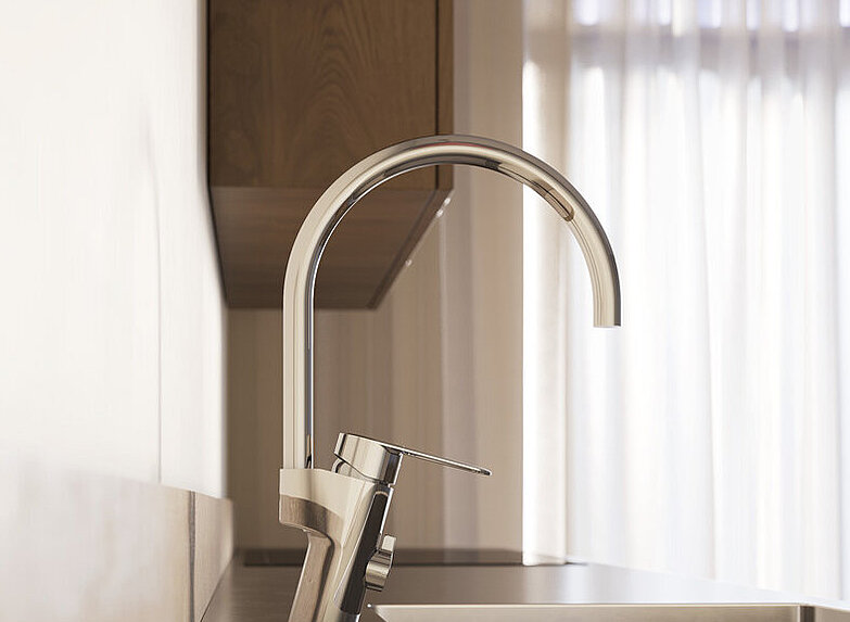 Mūsdienīgas virtuves izlietnes tuvplāns ar augstu hromētu virtuves jaucējkrānu un dabisko gaismu, kas ieplūst caur plāniem aizkariem fonā.