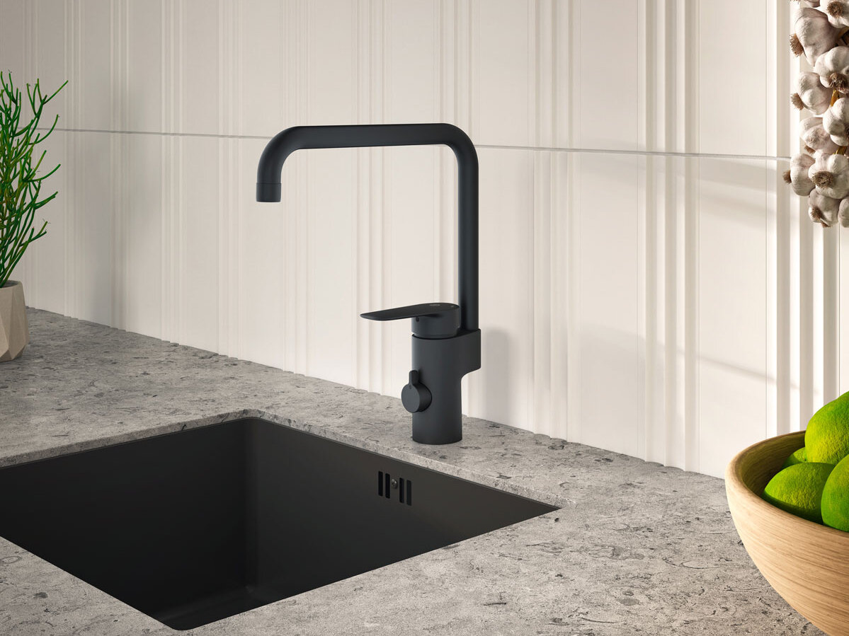En modern Gustavsberg svart köksblandare med smarta funktioner står över en snygg svart diskho på en grå bänkskiva. Bakstycket har vita vertikala linjer, kompletterat med en liten grön växt och en skål med limefrukter i närheten.