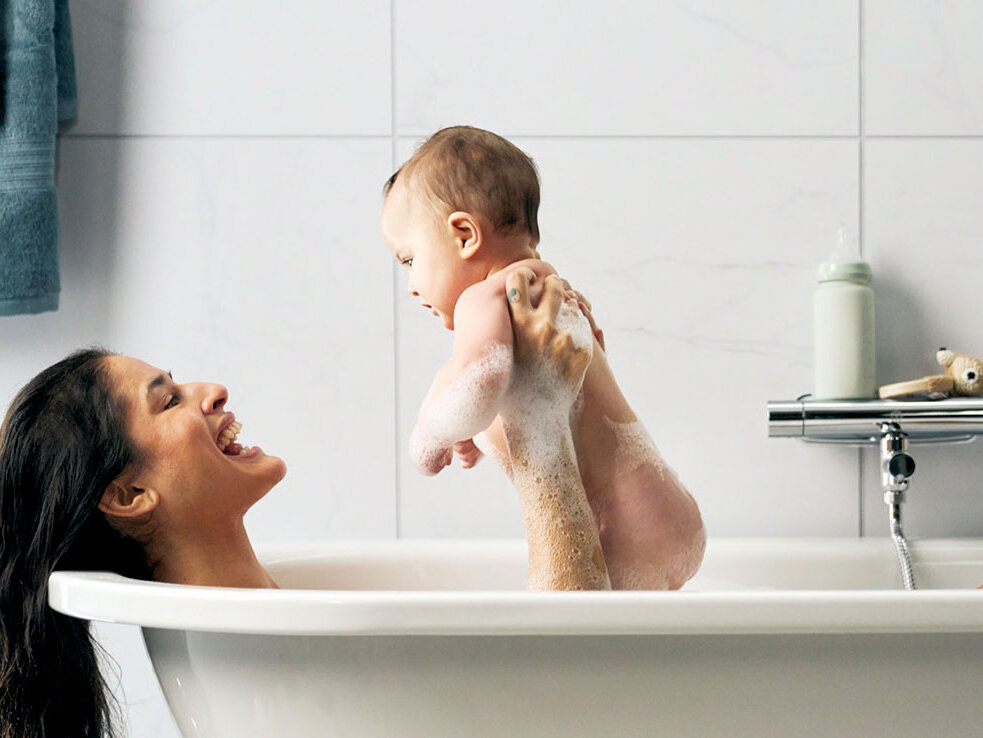 An adult holds a baby above the water while bathing together in a white bathtub in a tiled bathroom.