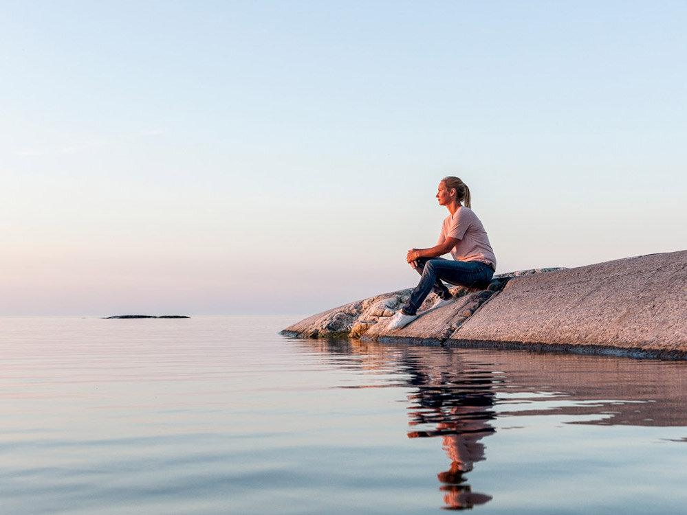 A person sits on a rock by calm water, looking out at the horizon during a clear, peaceful evening.