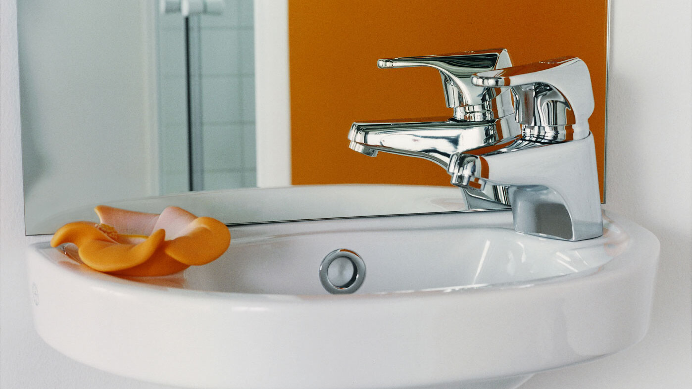 A white Gustavsberg sink with a chrome mixer, an orange flower, and a mirror reflecting part of an orange wall.