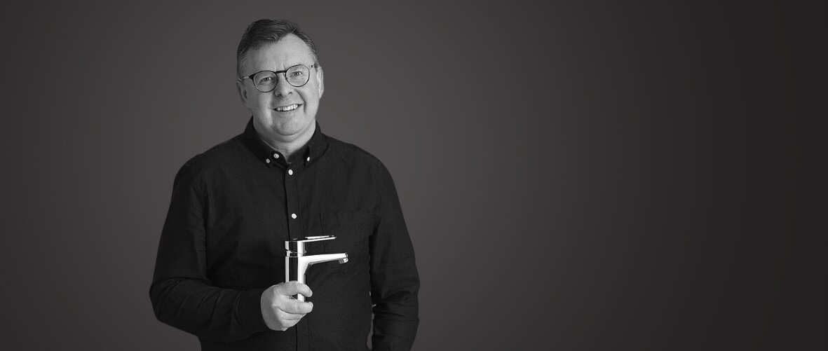 Mustapaitainen mies seisoo yksinkertaista tummaa taustaa vasten ja hymyilee kameralle yleiskonetta pitelemässä.