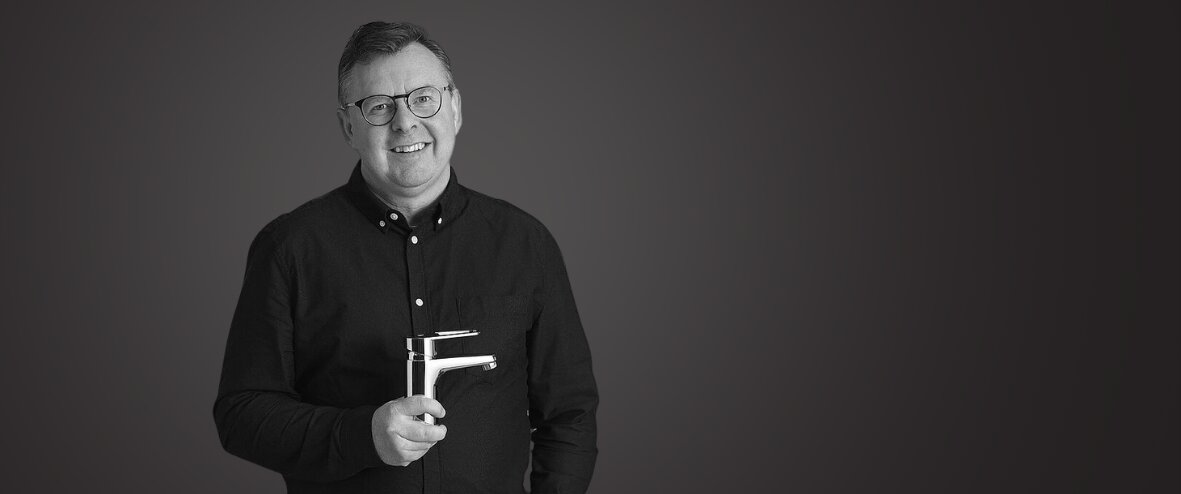 Mattias Virsgård i en sort skjorte, der holder en mixer, stående mod en almindelig mørk baggrund og smilende til kameraet.