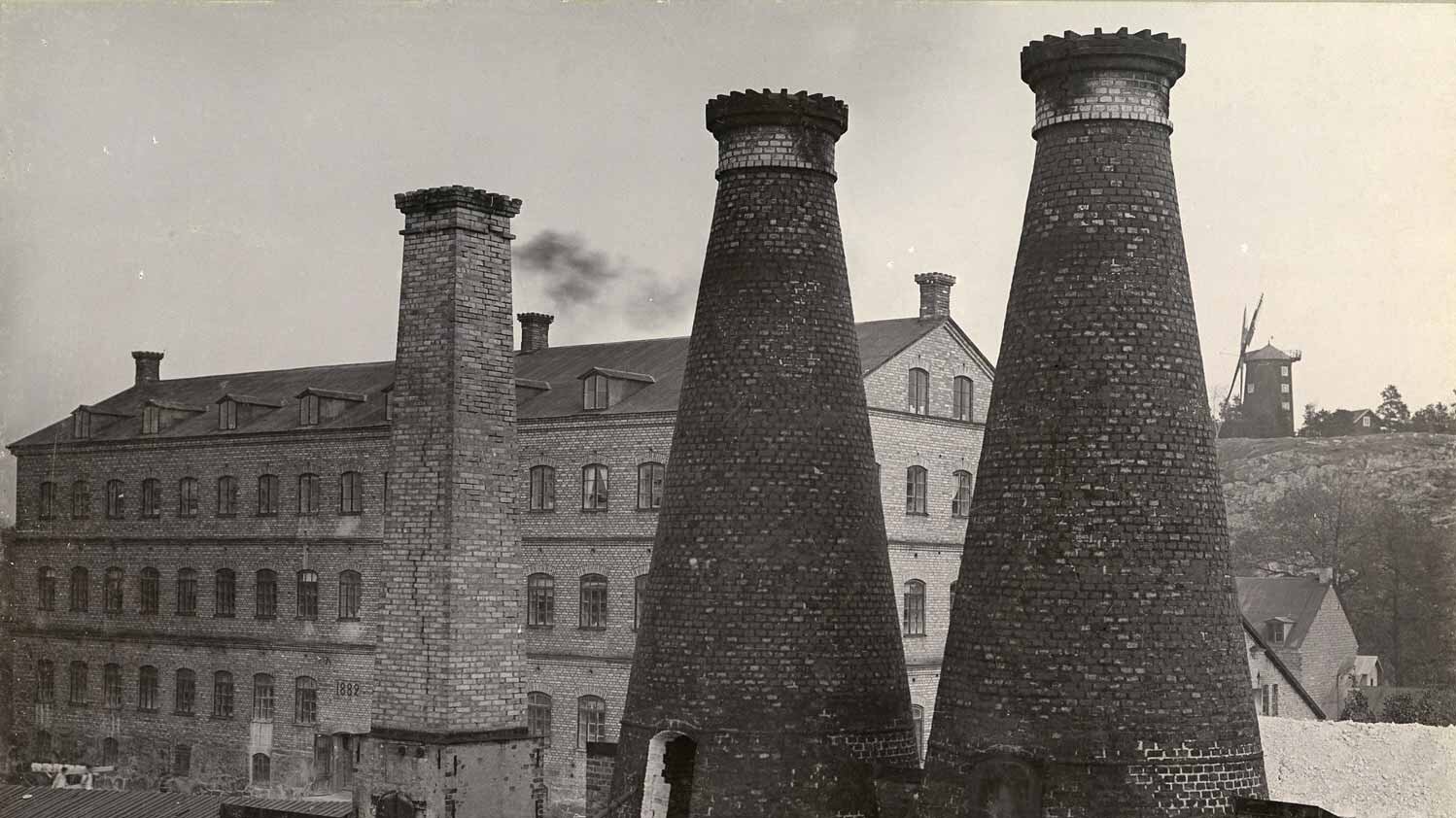 Black-and-white photo of Gustavsberg’s porcelain factory, chimneys rising near a windmill on a hill.