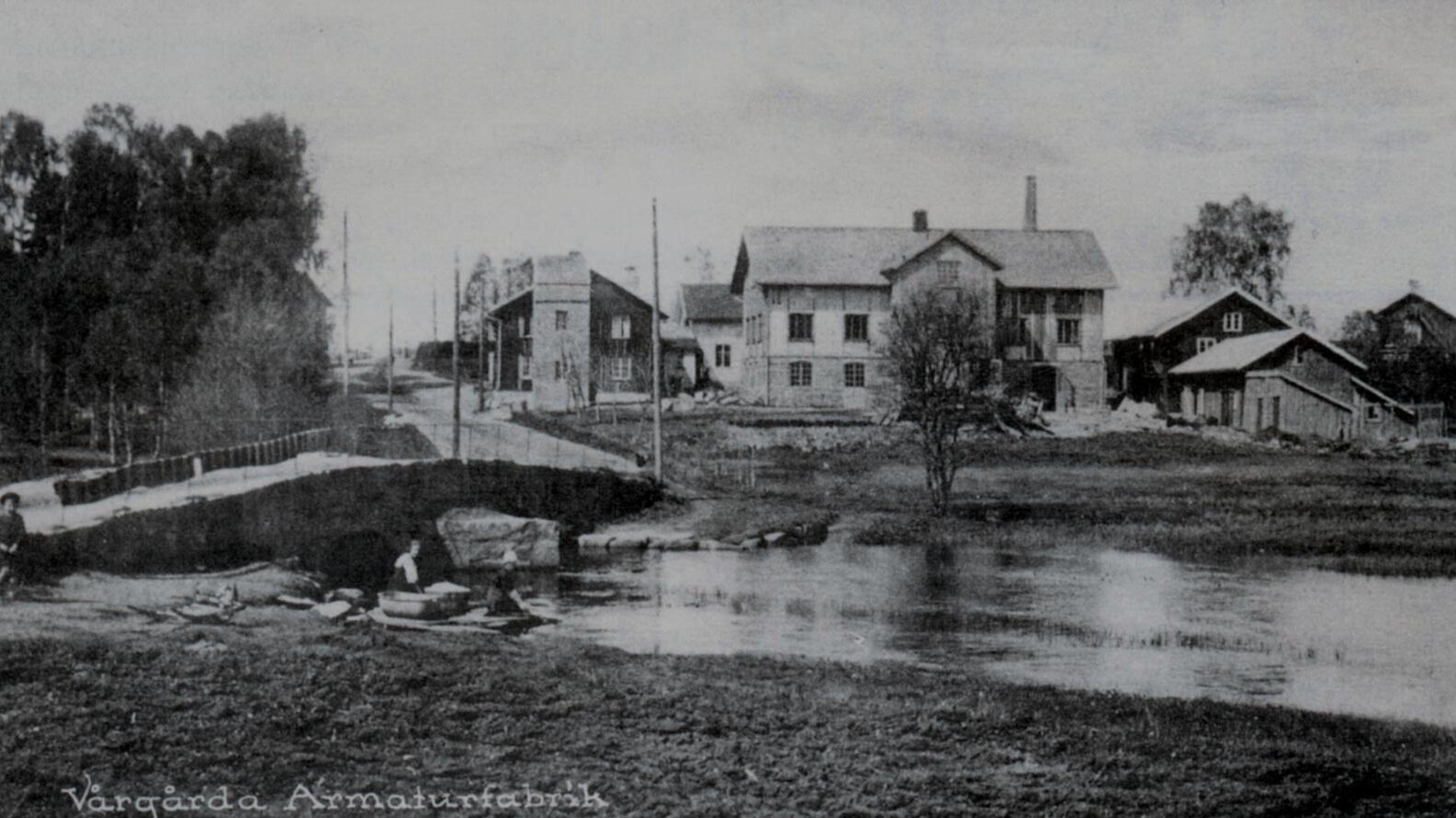 Black and white photo of the wooden buildings at the Vårgårda factory, located by a river with a small bridge.