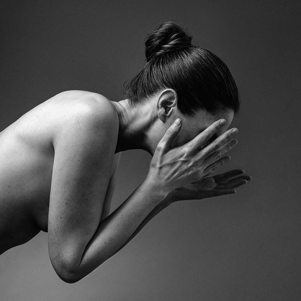 En kvinna med håret uppsatt i en knut tvättar ansiktet med båda händerna, fotograferad från sidan mot en enfärgad bakgrund.