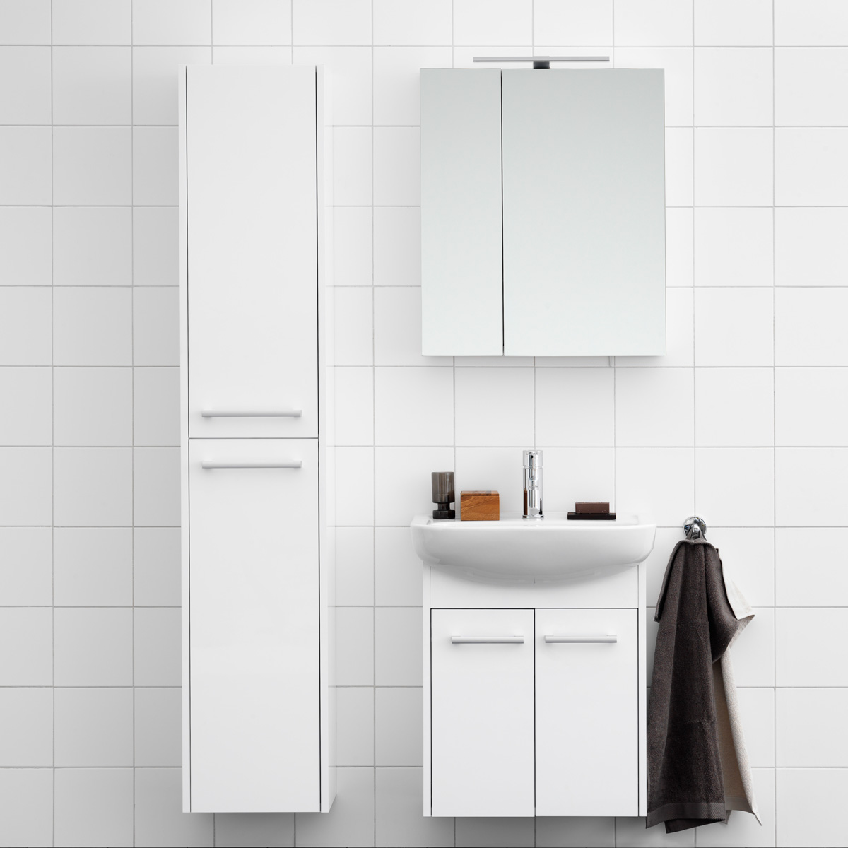 Minimalistisk hvitt bad med høyt skap, vask, speil og et mørkt håndkle hengende på en krok.