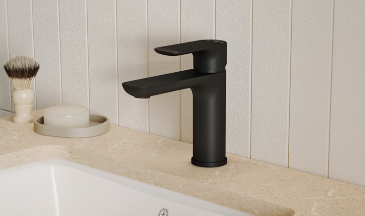 A black bathroom faucet on a beige stone countertop with a white sink and a shaving brush and soap next to it.